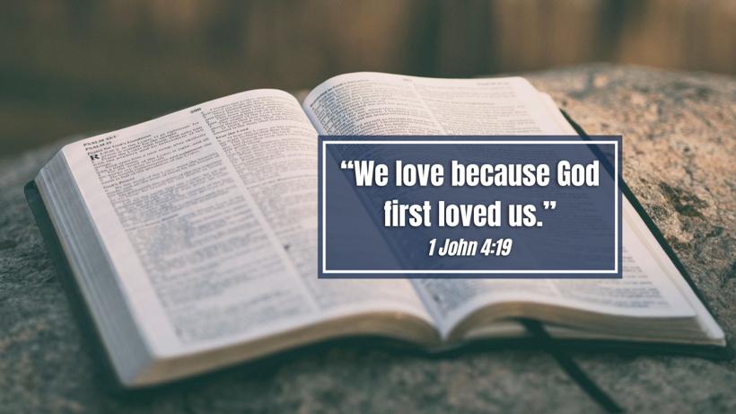 An open Bible on a stone ledge with the verse &ldquo;We love because God first loved us,&rdquo; 1 John 4:19